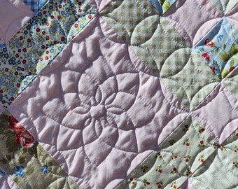 May include: A pink quilt with a floral pattern. The quilt is made of different fabrics, including gingham, floral prints, and solid colors. The quilt is quilted with a flower design in the center.