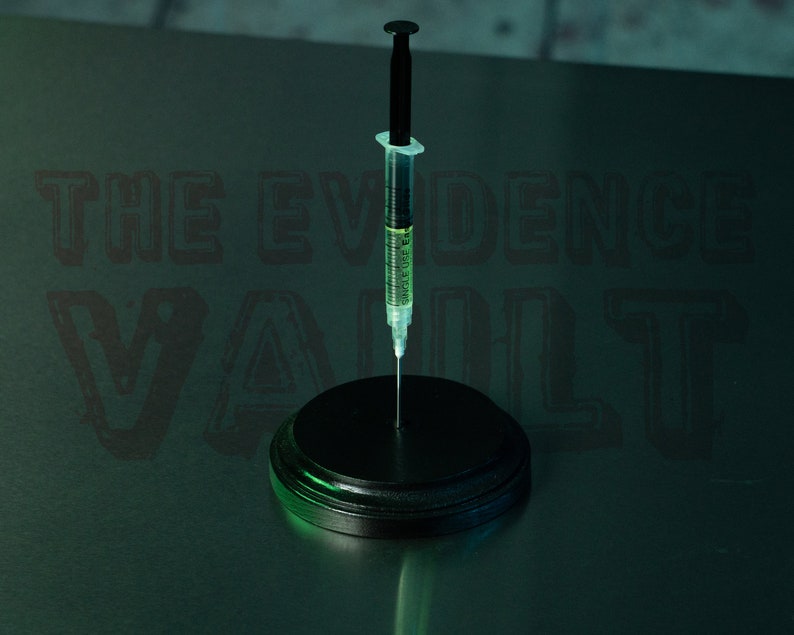 Puede incluir: Una jeringa negra con un l&iacute;quido verde en su interior est&aacute; de pie sobre una base circular negra. El fondo es de color verde oscuro con el texto "THE EVIDENCE VAULT" en negro.