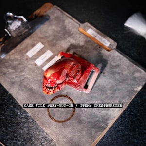May include: A prop chestburster alien model, with a red, fleshy appearance and sharp teeth, rests on a gray surface. The text "CASE FILE #WEY-YUT-CB / ITEM: CHESTBURSTER" is visible. A metal clipboard and other props are in the background.