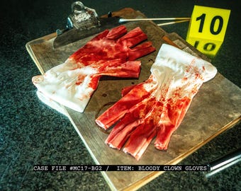 May include: A pair of white clown gloves heavily stained with red, resembling blood, rest on a wooden clipboard. A yellow evidence marker with the number 10 is visible. Text on the image reads: CASE FILE #MC17-BG2 / ITEM: BLOODY CLOWN GLOVES.