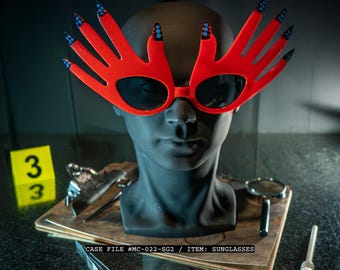 May include: Red sunglasses in the shape of hands with black lenses and blue and black nail accents. The sunglasses are displayed on a black mannequin head. The text on the image reads "CASE FILE #MC-022-SG2 / ITEM: SUNGLASSES."