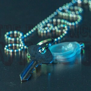 May include: A silver key chain with a silver key and a clear plastic charm. The key chain is lying on a dark blue surface.