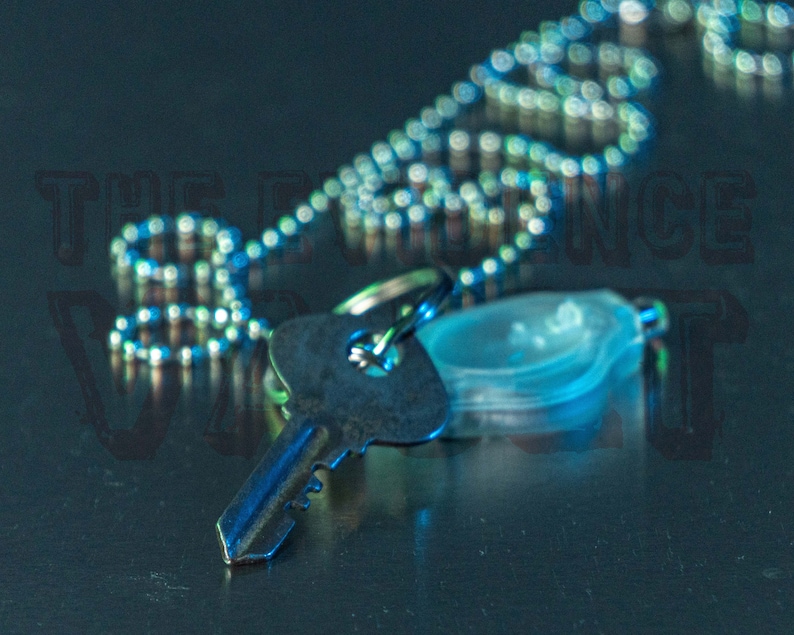 May include: A silver key chain with a silver key and a clear plastic charm. The key chain is lying on a dark blue surface.