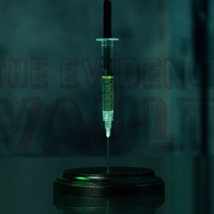 Puede incluir: Una jeringa de vidrio transparente llena de un l&iacute;quido verde est&aacute; montada sobre una base circular negra. La jeringa est&aacute; colocada frente a un fondo verde oscuro con el texto "THE EVIDENCE VAULT" en blanco.