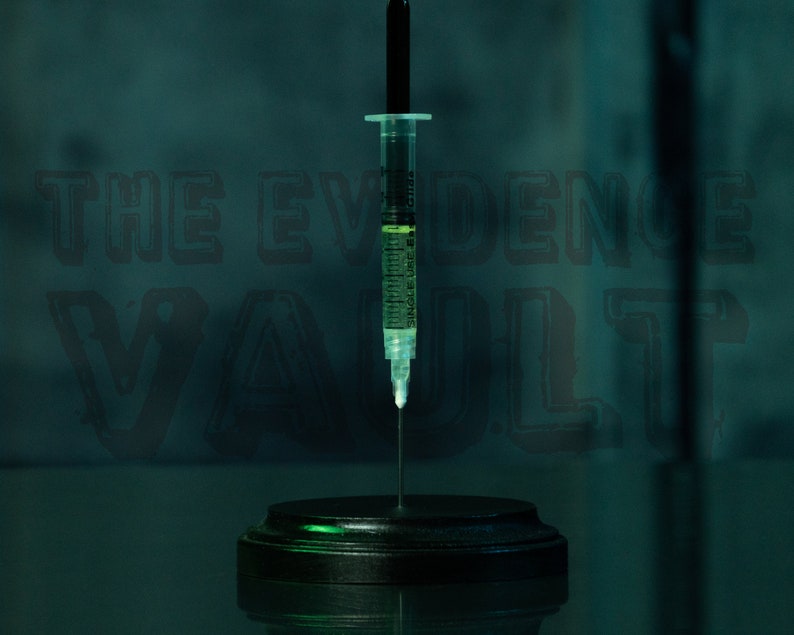 Puede incluir: Una jeringa de vidrio transparente llena de un l&iacute;quido verde est&aacute; montada sobre una base circular negra. La jeringa est&aacute; colocada frente a un fondo verde oscuro con el texto "THE EVIDENCE VAULT" en blanco.