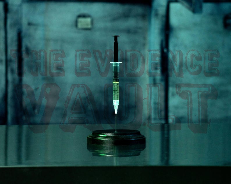 Puede incluir: Una jeringa de vidrio transparente llena de un l&iacute;quido amarillo se encuentra sobre un soporte circular negro. La jeringa est&aacute; delante de una pared azul con el texto "THE EVIDENCE VAULT" escrito en negro.