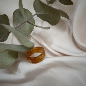Könnte beinhalten: Ein brauner Harzring mit Marmormuster. Der Ring liegt auf einem weißen Stoffhintergrund.