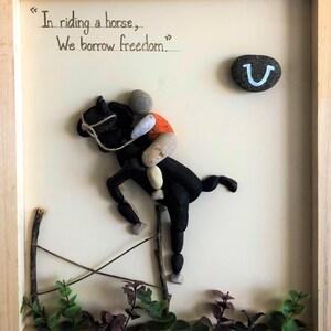 May include: A framed artwork featuring a horse and rider made from small stones. The horse is black and the rider is wearing an orange shirt. The artwork includes a horseshoe and the quote "In riding a horse, we borrow freedom."