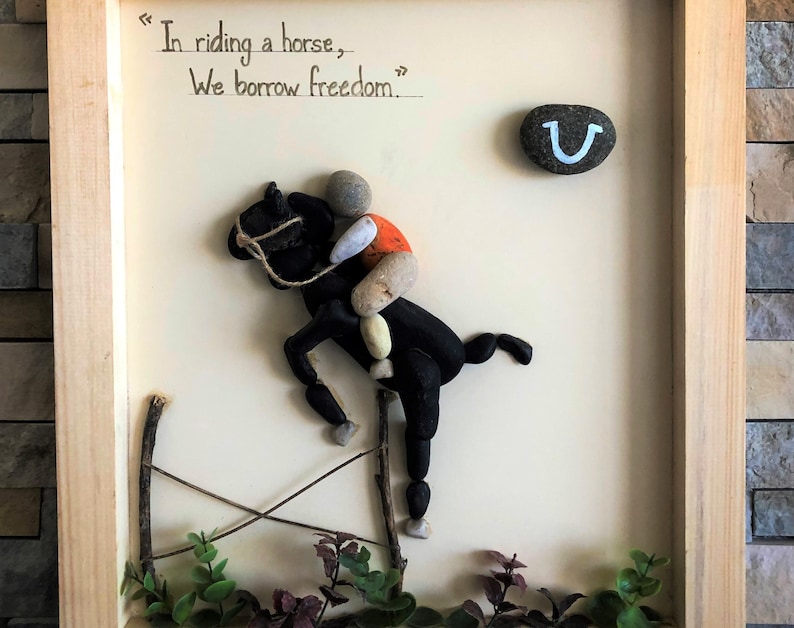 May include: A framed artwork featuring a horse and rider made from small stones. The horse is black and the rider is wearing an orange shirt. The artwork includes a horseshoe and the quote "In riding a horse, we borrow freedom."