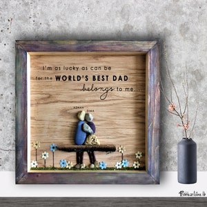 May include: Wooden framed artwork featuring a pebble art design of two figures sitting on a bench. The artwork includes the text "I'm as lucky as can be for the WORLD'S BEST DAD belongs to me." and the names "ADNAN" and "DIMA" written below the figures.