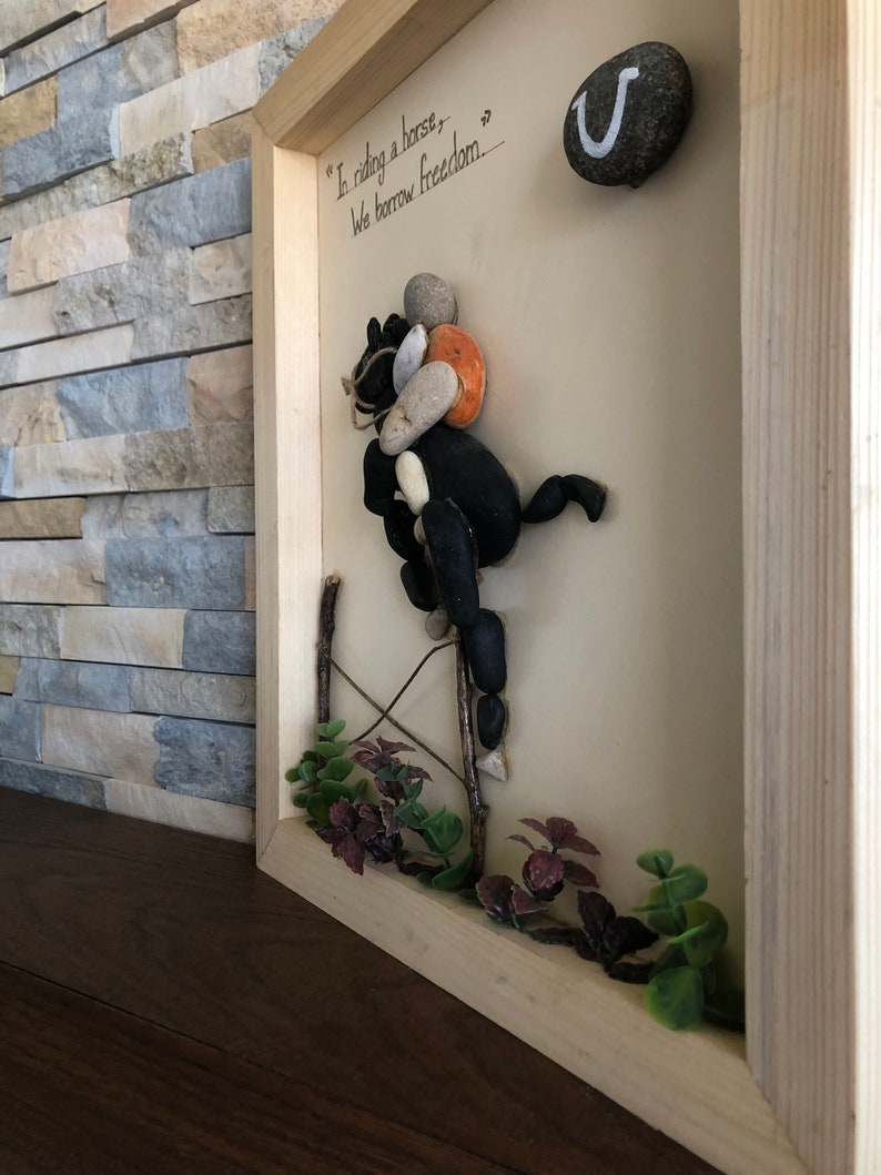 May include: A framed artwork made with stones depicting a horse and rider with the quote "In riding a horse, we borrow freedom." A horseshoe is in the upper right corner.