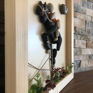 May include: A framed artwork made with small stones, twigs, and greenery. The artwork depicts a black horse with a rider, with the text "Your freedom" written on the white background.