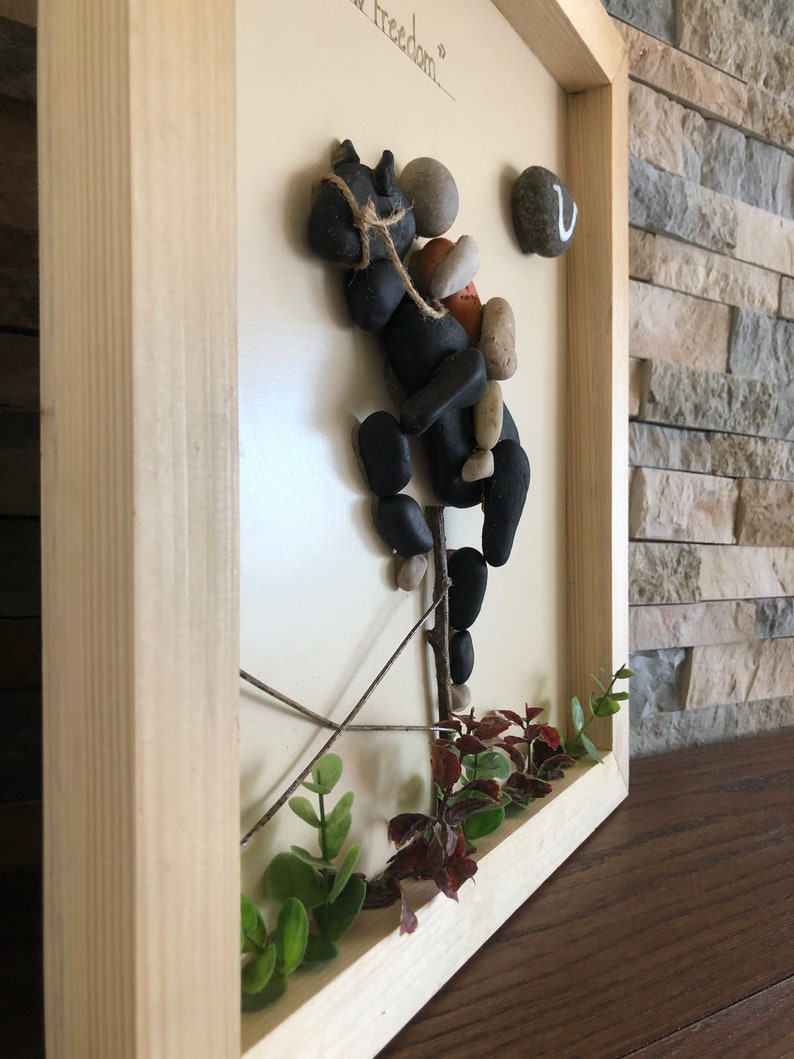 May include: A framed artwork made with small stones, twigs, and greenery. The artwork depicts a black horse with a rider, with the text "Your freedom" written on the white background.