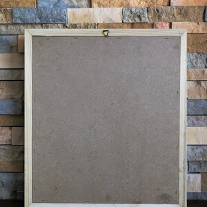 May include: A wooden picture frame with a brown cardboard backing. The frame is hanging on a wall made of stacked stone tiles.