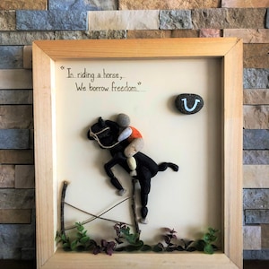 May include: A framed artwork featuring a horse and rider made of stones, with the quote "In riding a horse, we borrow freedom." The artwork also includes a horseshoe with a smiley face.
