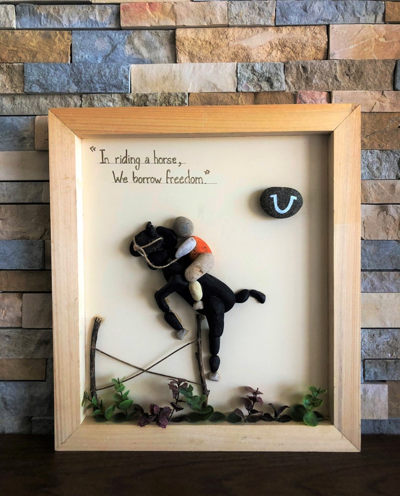 May include: A framed artwork featuring a horse and rider made of stones, with the quote "In riding a horse, we borrow freedom." The artwork also includes a horseshoe with a smiley face.