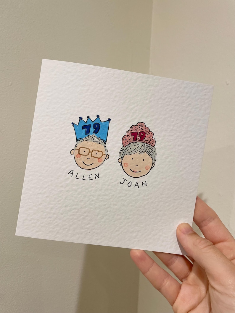 Puede incluir: Tarjeta de felicitaci&oacute;n dibujada a mano con dos caras de dibujos animados, cada una con una corona con el n&uacute;mero 79. Los nombres "Allen" y "Joan" est&aacute;n escritos debajo. La tarjeta es blanca con una superficie texturizada.
