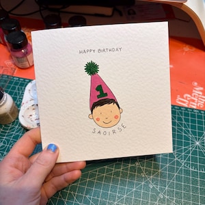 Puede incluir: Tarjeta de cumplea&ntilde;os cuadrada con el texto "HAPPY BIRTHDAY". La tarjeta presenta una ilustraci&oacute;n de un ni&ntilde;o con un gorro de fiesta rosa con el n&uacute;mero "1" y el nombre "SAOIRSE". El gorro tiene un pomp&oacute;n verde.