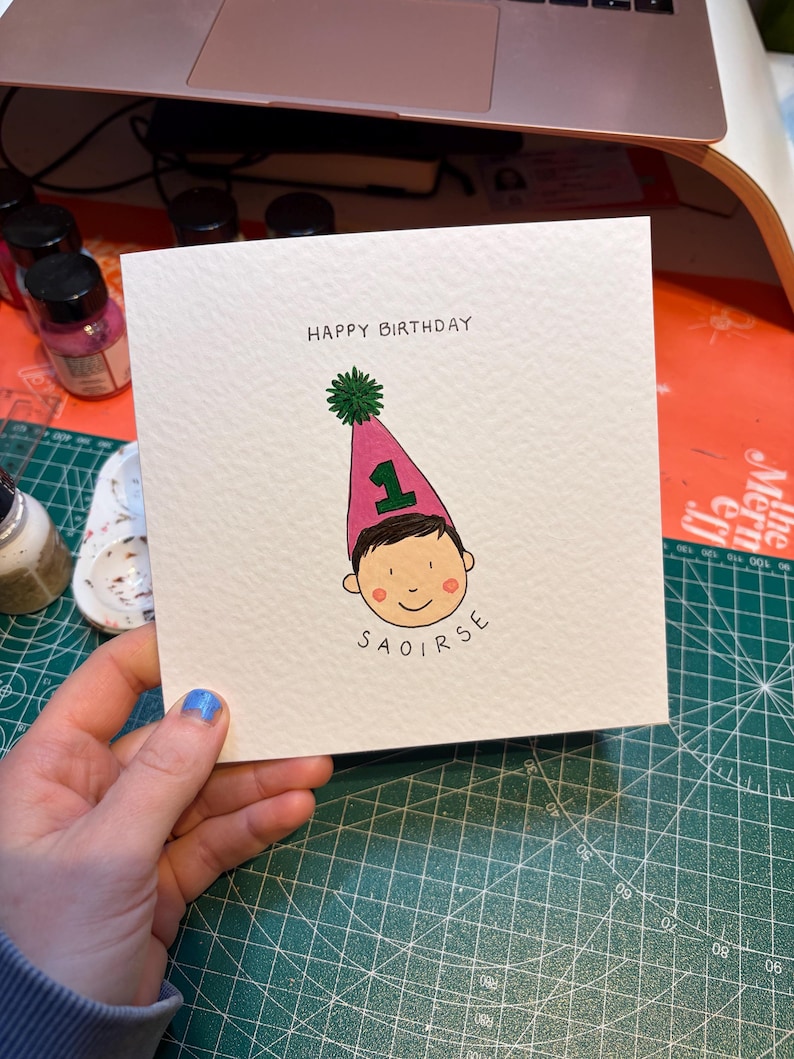 Puede incluir: Tarjeta de cumplea&ntilde;os cuadrada con el texto "HAPPY BIRTHDAY". La tarjeta presenta una ilustraci&oacute;n de un ni&ntilde;o con un gorro de fiesta rosa con el n&uacute;mero "1" y el nombre "SAOIRSE". El gorro tiene un pomp&oacute;n verde.