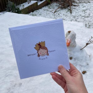 Puede incluir: Una tarjeta de cumplea&ntilde;os blanca con el texto "Birthday Queen Minnie" y una ilustraci&oacute;n de una ni&ntilde;a con una corona. La tarjeta se sostiene frente a un fondo nevado con un mu&ntilde;eco de nieve.