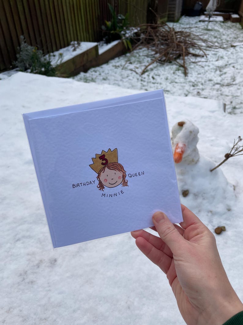Puede incluir: Una tarjeta de cumplea&ntilde;os blanca con el texto "Birthday Queen Minnie" y una ilustraci&oacute;n de una ni&ntilde;a con una corona. La tarjeta se sostiene frente a un fondo nevado con un mu&ntilde;eco de nieve.