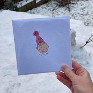 Puede incluir: Tarjeta de felicitaci&oacute;n cuadrada con una ilustraci&oacute;n de dibujos animados de un beb&eacute; con un gorro de fiesta rosa con el n&uacute;mero 1. El beb&eacute; tiene una cara sonriente y la palabra "FLISS" est&aacute; escrita debajo. La tarjeta est&aacute; en una funda de pl&aacute;stico transparente.
