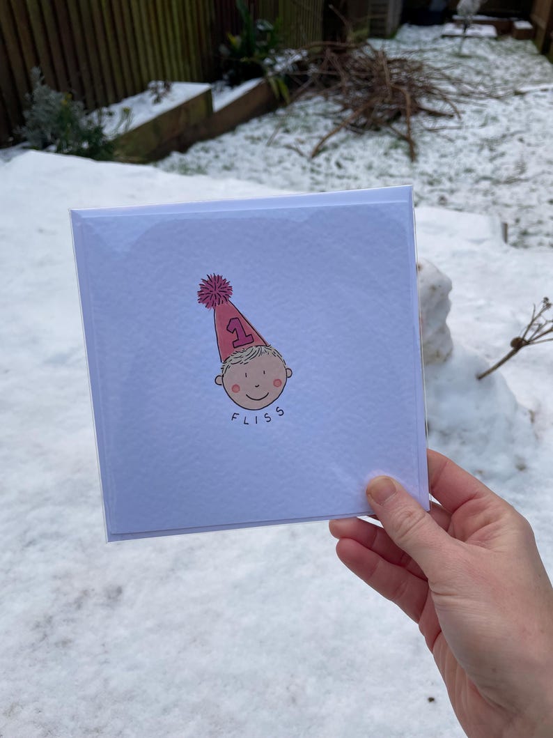 Puede incluir: Tarjeta de felicitaci&oacute;n cuadrada con una ilustraci&oacute;n de dibujos animados de un beb&eacute; con un gorro de fiesta rosa con el n&uacute;mero 1. El beb&eacute; tiene una cara sonriente y la palabra "FLISS" est&aacute; escrita debajo. La tarjeta est&aacute; en una funda de pl&aacute;stico transparente.