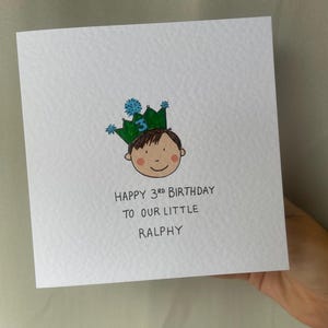 Puede incluir: Una tarjeta de felicitaci&oacute;n cuadrada blanca con una ilustraci&oacute;n dibujada a mano de un ni&ntilde;o con una corona verde con el n&uacute;mero 3. El texto dice "HAPPY 3RD BIRTHDAY TO OUR LITTLE RALPHY".