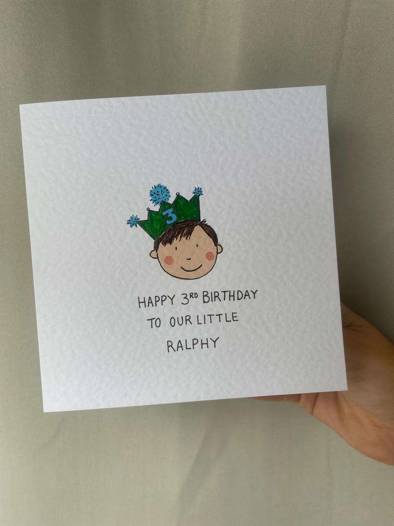 Puede incluir: Una tarjeta de felicitaci&oacute;n cuadrada blanca con una ilustraci&oacute;n dibujada a mano de un ni&ntilde;o con una corona verde con el n&uacute;mero 3. El texto dice "HAPPY 3RD BIRTHDAY TO OUR LITTLE RALPHY".