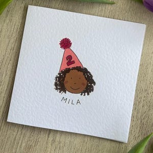 Puede incluir: Tarjeta de cumplea&ntilde;os con una ilustraci&oacute;n de dibujos animados de un ni&ntilde;o con piel morena y cabello rizado. El ni&ntilde;o lleva un gorro de fiesta rosa con el n&uacute;mero 2. El nombre "MILA" est&aacute; escrito debajo.