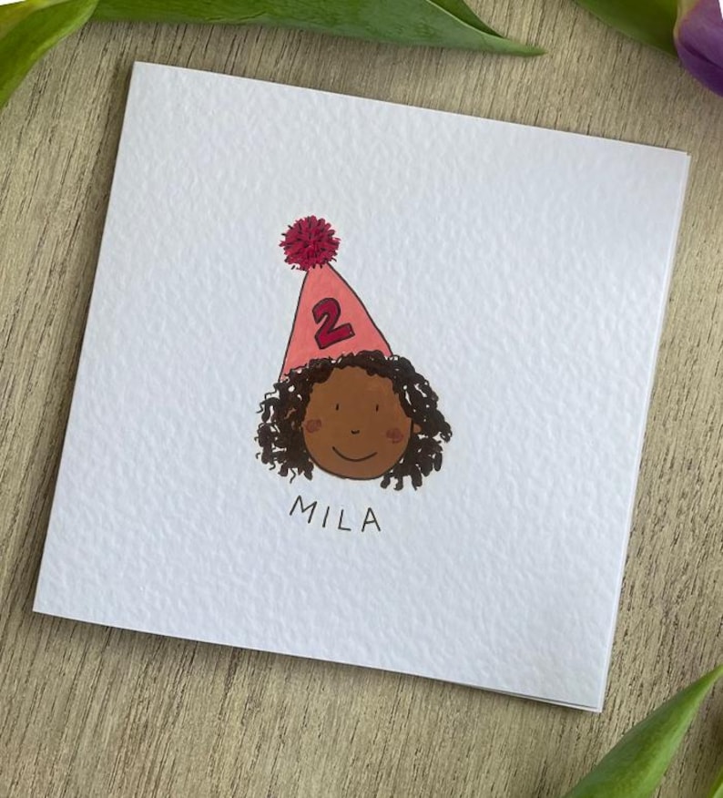 Puede incluir: Tarjeta de cumplea&ntilde;os con una ilustraci&oacute;n de dibujos animados de un ni&ntilde;o con piel morena y cabello rizado. El ni&ntilde;o lleva un gorro de fiesta rosa con el n&uacute;mero 2. El nombre "MILA" est&aacute; escrito debajo.