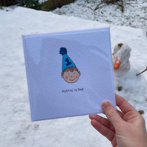 Puede incluir: Una tarjeta de felicitaci&oacute;n cuadrada con el texto "AUSTIN IS ONE" presenta una ilustraci&oacute;n de dibujos animados de un ni&ntilde;o con un gorro de fiesta azul con el n&uacute;mero 1. La tarjeta se sostiene frente a un fondo nevado con un mu&ntilde;eco de nieve.