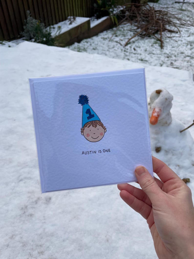 Puede incluir: Una tarjeta de felicitaci&oacute;n cuadrada con el texto "AUSTIN IS ONE" presenta una ilustraci&oacute;n de dibujos animados de un ni&ntilde;o con un gorro de fiesta azul con el n&uacute;mero 1. La tarjeta se sostiene frente a un fondo nevado con un mu&ntilde;eco de nieve.