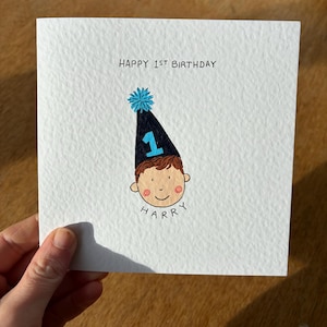 Puede incluir: Tarjeta blanca con el texto "HAPPY 1ST BIRTHDAY" y una ilustraci&oacute;n de dibujos animados de la cara de un ni&ntilde;o con un gorro de fiesta azul y negro con el n&uacute;mero 1. El nombre "HARRY" est&aacute; escrito debajo.
