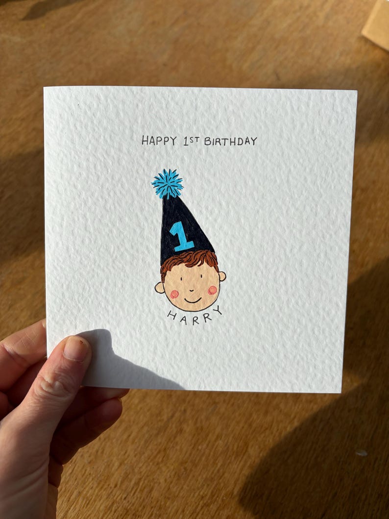 Puede incluir: Tarjeta blanca con el texto "HAPPY 1ST BIRTHDAY" y una ilustraci&oacute;n de dibujos animados de la cara de un ni&ntilde;o con un gorro de fiesta azul y negro con el n&uacute;mero 1. El nombre "HARRY" est&aacute; escrito debajo.