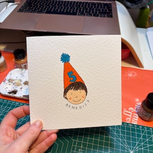 Puede incluir: Tarjeta de felicitaci&oacute;n cuadrada con una ilustraci&oacute;n de dibujos animados de un ni&ntilde;o con un gorro de cumplea&ntilde;os con el n&uacute;mero 5. El nombre del ni&ntilde;o, "Benedict", est&aacute; escrito debajo de la cara. El sombrero es naranja con un pomp&oacute;n azul.