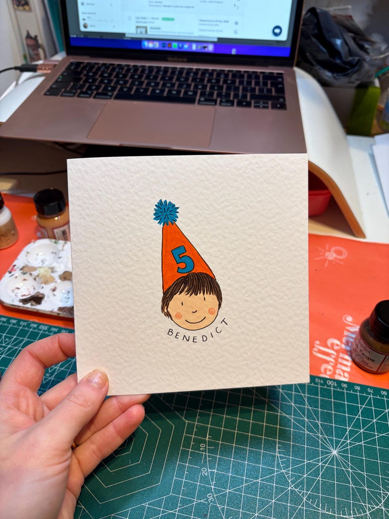 Puede incluir: Tarjeta de felicitaci&oacute;n cuadrada con una ilustraci&oacute;n de dibujos animados de un ni&ntilde;o con un gorro de cumplea&ntilde;os con el n&uacute;mero 5. El nombre del ni&ntilde;o, "Benedict", est&aacute; escrito debajo de la cara. El sombrero es naranja con un pomp&oacute;n azul.