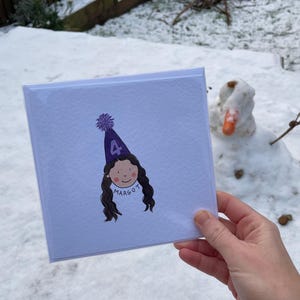 Puede incluir: Una tarjeta de felicitaci&oacute;n cuadrada con una ilustraci&oacute;n de dibujos animados de una ni&ntilde;a con un gorro de fiesta morado con el n&uacute;mero 4. La ni&ntilde;a tiene el pelo largo y casta&ntilde;o y el nombre "Margot" est&aacute; escrito debajo. La tarjeta se sostiene frente a un fondo nevado.