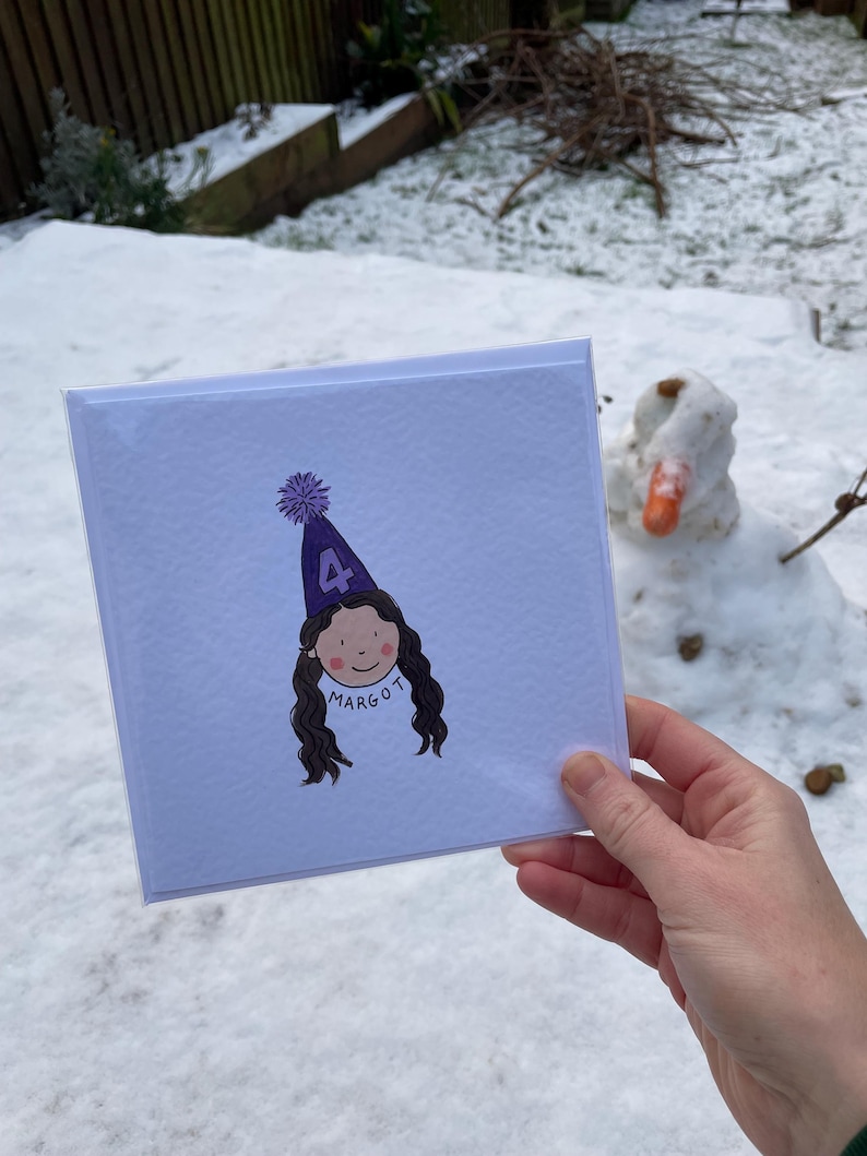 Puede incluir: Una tarjeta de felicitaci&oacute;n cuadrada con una ilustraci&oacute;n de dibujos animados de una ni&ntilde;a con un gorro de fiesta morado con el n&uacute;mero 4. La ni&ntilde;a tiene el pelo largo y casta&ntilde;o y el nombre "Margot" est&aacute; escrito debajo. La tarjeta se sostiene frente a un fondo nevado.