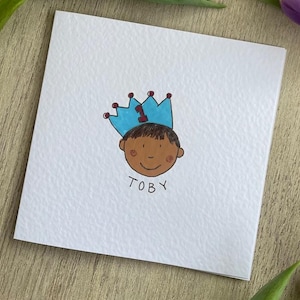 Puede incluir: Una tarjeta blanca con una ilustraci&oacute;n dibujada a mano de un ni&ntilde;o con una corona azul con el n&uacute;mero 1. El ni&ntilde;o tiene la piel morena y el nombre "TOBY" est&aacute; escrito debajo. La tarjeta est&aacute; sobre una superficie de madera con hojas verdes.