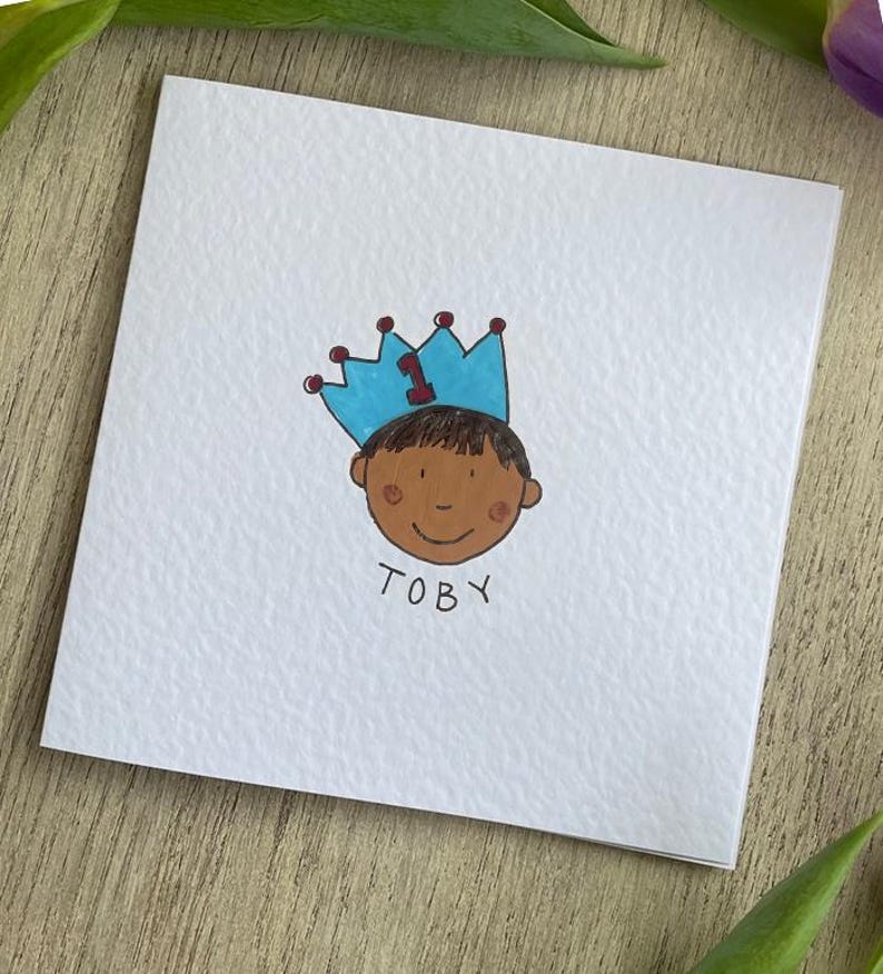 Puede incluir: Una tarjeta blanca con una ilustraci&oacute;n dibujada a mano de un ni&ntilde;o con una corona azul con el n&uacute;mero 1. El ni&ntilde;o tiene la piel morena y el nombre "TOBY" est&aacute; escrito debajo. La tarjeta est&aacute; sobre una superficie de madera con hojas verdes.