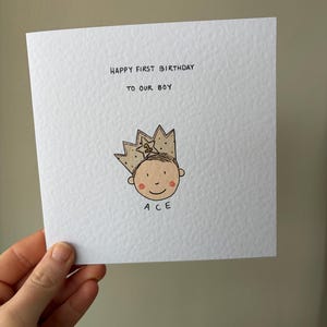 Puede incluir: Una tarjeta de felicitaci&oacute;n cuadrada blanca con el texto "HAPPY FIRST BIRTHDAY TO OUR BOY". Debajo hay una ilustraci&oacute;n de dibujos animados de una cara sonriente con una corona dorada. El nombre "ACE" est&aacute; escrito debajo.