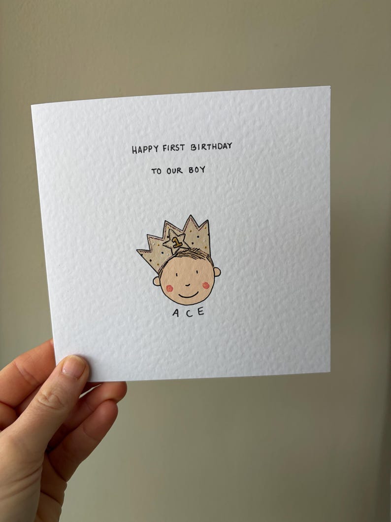 Puede incluir: Una tarjeta de felicitaci&oacute;n cuadrada blanca con el texto "HAPPY FIRST BIRTHDAY TO OUR BOY". Debajo hay una ilustraci&oacute;n de dibujos animados de una cara sonriente con una corona dorada. El nombre "ACE" est&aacute; escrito debajo.
