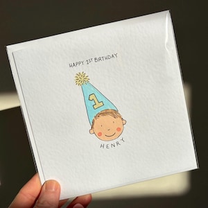 Puede incluir: Tarjeta de felicitaci&oacute;n blanca con el texto "HAPPY 1ST BIRTHAY" y el nombre "HENRY". La tarjeta presenta una ilustraci&oacute;n de dibujos animados de la cara de un ni&ntilde;o con un gorro de fiesta azul con una estrella amarilla.