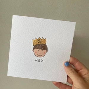 Puede incluir: Una tarjeta cuadrada blanca con una ilustraci&oacute;n dibujada a mano de la cara de un ni&ntilde;o con una corona dorada con el n&uacute;mero 3. El ni&ntilde;o tiene cabello casta&ntilde;o y una cara sonriente. El nombre "REX" est&aacute; escrito debajo.