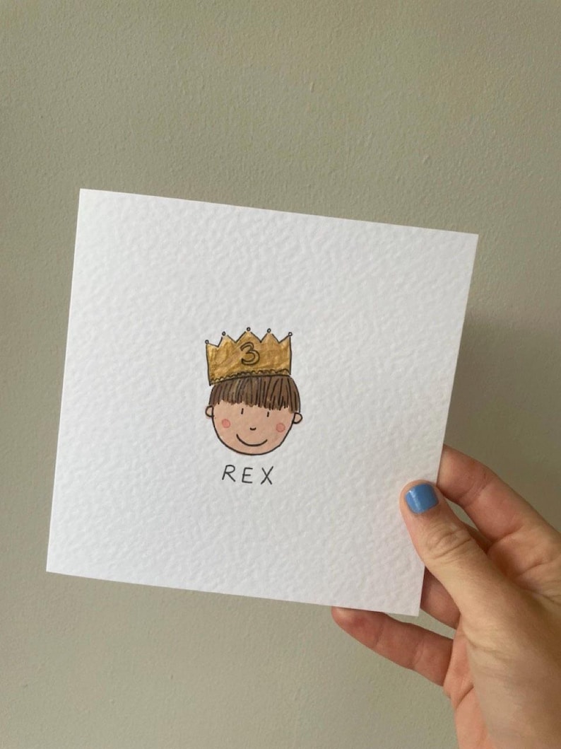 Puede incluir: Una tarjeta cuadrada blanca con una ilustraci&oacute;n dibujada a mano de la cara de un ni&ntilde;o con una corona dorada con el n&uacute;mero 3. El ni&ntilde;o tiene cabello casta&ntilde;o y una cara sonriente. El nombre "REX" est&aacute; escrito debajo.