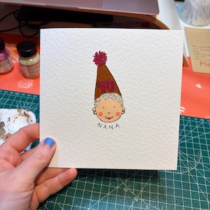 Puede incluir: Tarjeta de felicitaci&oacute;n cuadrada con una ilustraci&oacute;n dibujada a mano de una cara de dibujos animados con un gorro de fiesta marr&oacute;n con el n&uacute;mero 70. La palabra "NANA" est&aacute; escrita debajo. La tarjeta est&aacute; sobre una esterilla de corte verde.