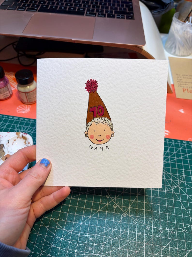 Puede incluir: Tarjeta de felicitaci&oacute;n cuadrada con una ilustraci&oacute;n dibujada a mano de una cara de dibujos animados con un gorro de fiesta marr&oacute;n con el n&uacute;mero 70. La palabra "NANA" est&aacute; escrita debajo. La tarjeta est&aacute; sobre una esterilla de corte verde.