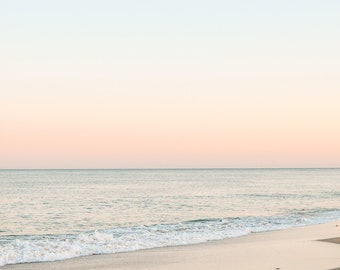May include: A serene coastal scene featuring a gradient sky transitioning from light blue to soft pink, meeting the calm ocean. Gentle waves lap the sandy shore, creating a peaceful, minimalist landscape.