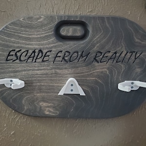 May include: A dark wood wall mount with the text "ESCAPE FROM REALITY" carved into it. The mount has three white plastic hooks for hanging VR headsets.
